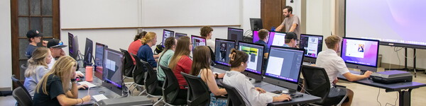 Wide view of the multi-media classroom with several large computer monitors with students working on projects.