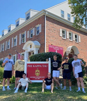 Young men standing and sitting around the Phi Kappa Tau sign in front of their house.