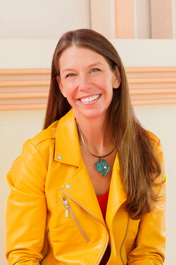Deb Mashek, a woman with long brown hair, smiles at the camera. She is wearing an orange blazer and is situated in front of a white and orange background.