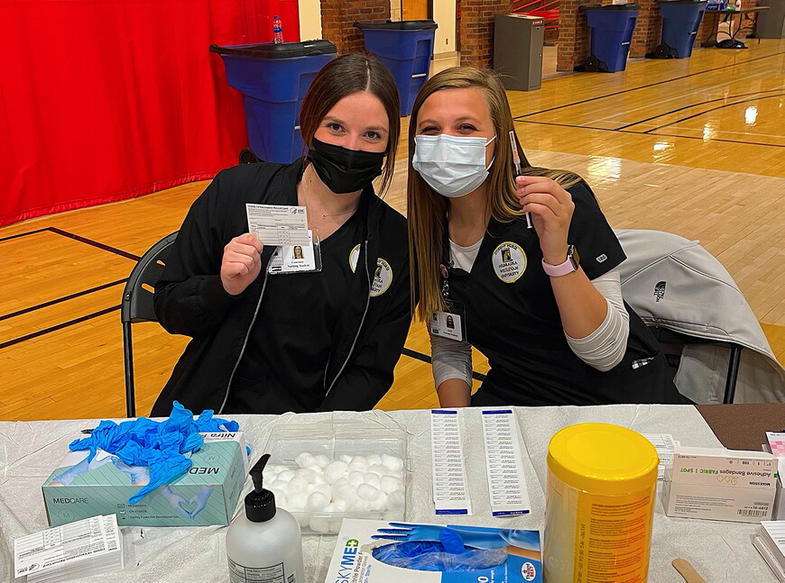 Two nursing students sitting in front of a table with vaccination supplies.