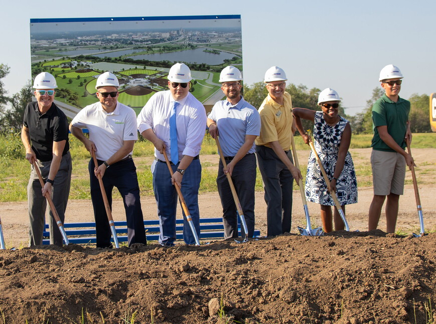 Community leaders wearing hard hats and holding shovels break ground at the new sports complex .