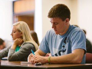 Male student in the foreground at a desk writing. Students at desks in the background.