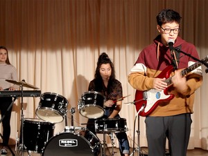 A student playing guitar, a student playing the drums and a student at playing a keyboard.