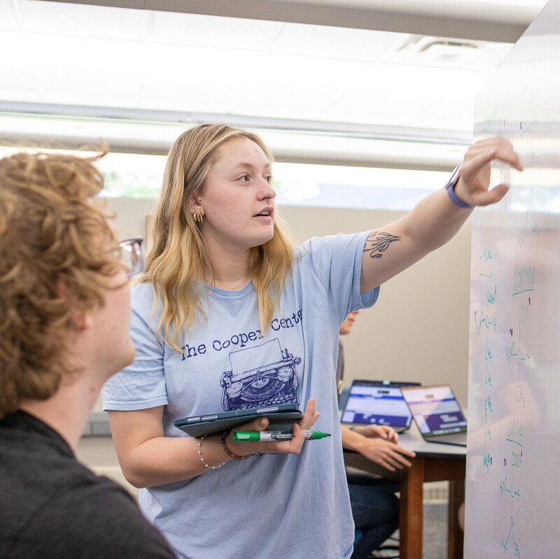 A female student standing in front of a whiteboard point to a formula as a seated male student looks on.