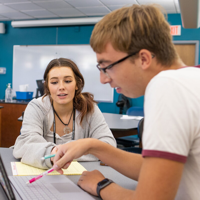 A female and male student seated at a table looking at a laptop screen.
