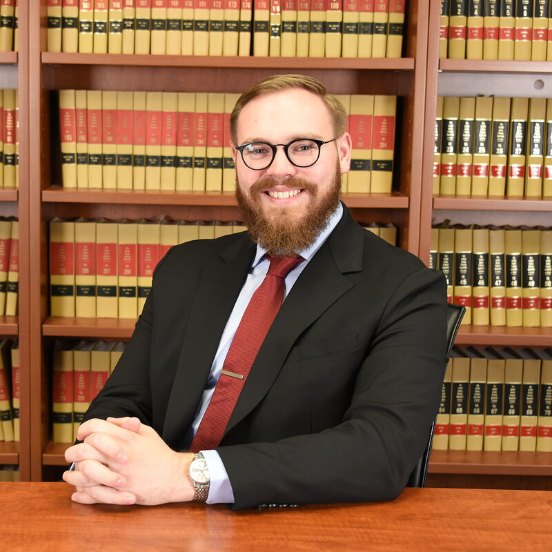 Adam Braegelman sitting with his hands folded on a table in front of a bookcase.