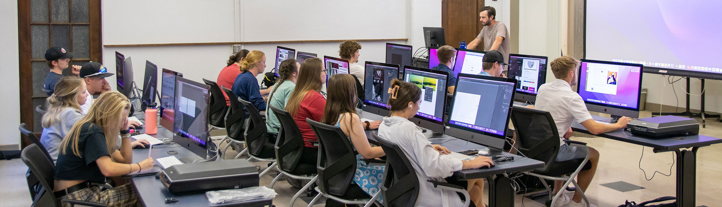 Classroom with three rows of desks with computers with big monitors. Students are seated at desks working with an instructor at the front of the class.