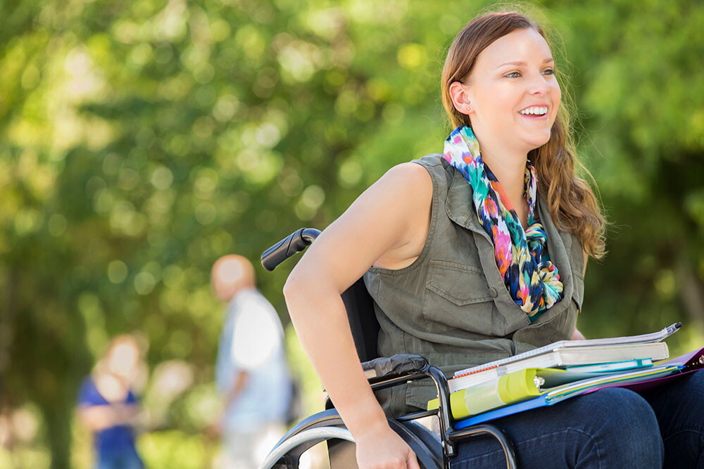Young woman in wheel chair with books on her lap, outside on a nice day.
