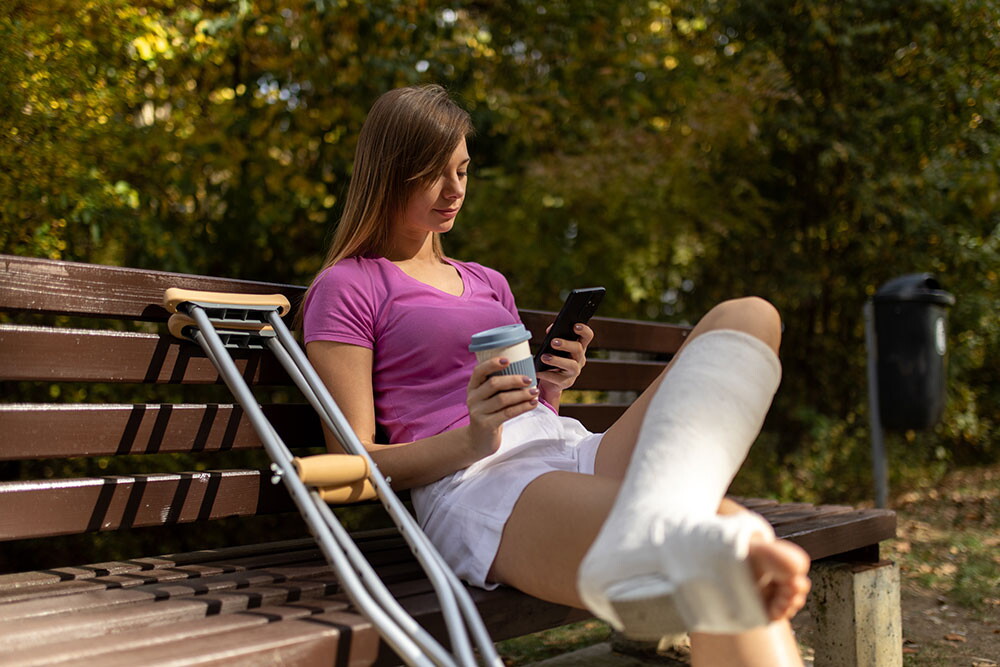 Woman with a leg cast and crutches sits on a bench holding a cup and phone.