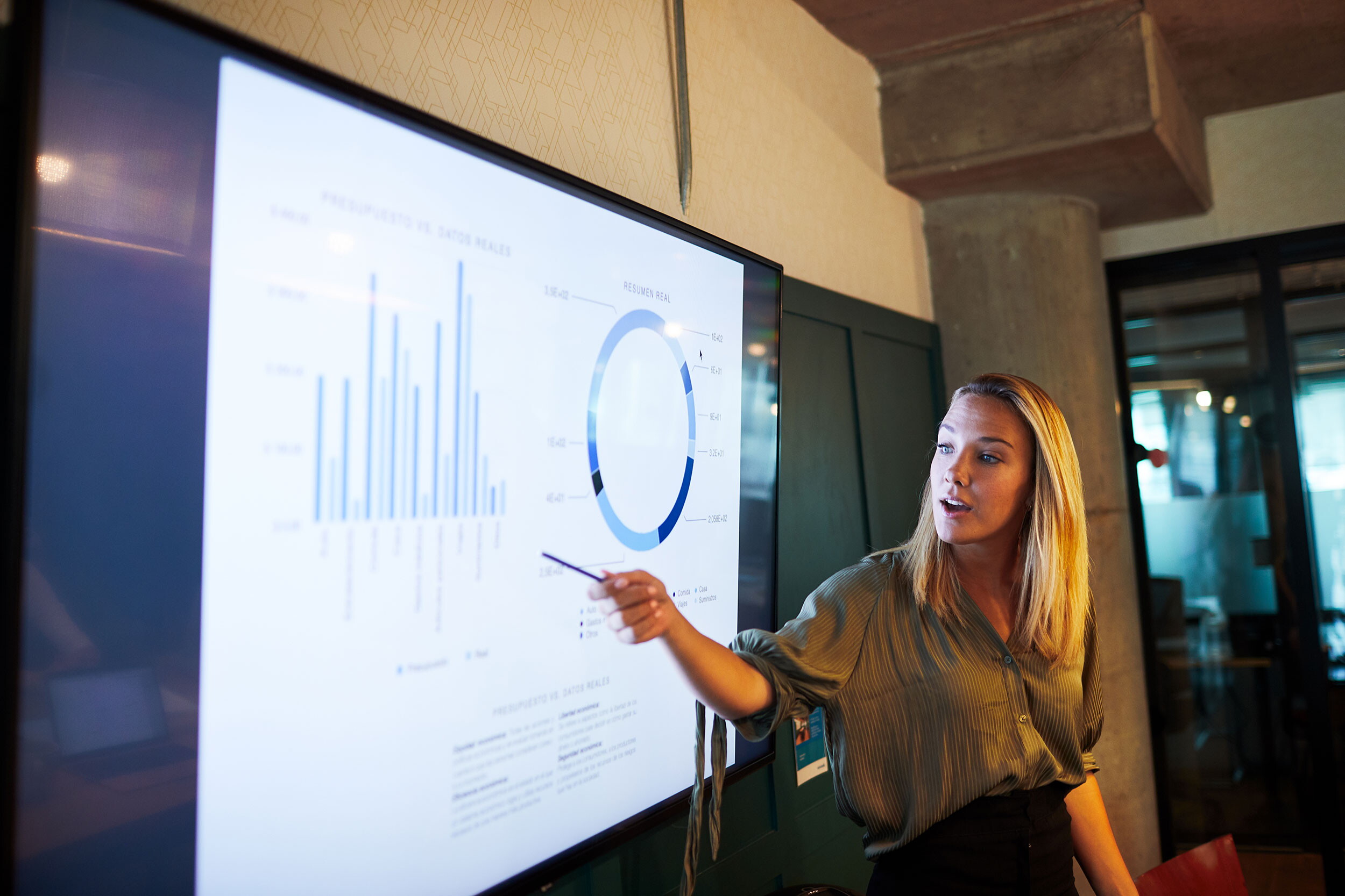 Woman stands and points to a graph on a large monitor while speaking.