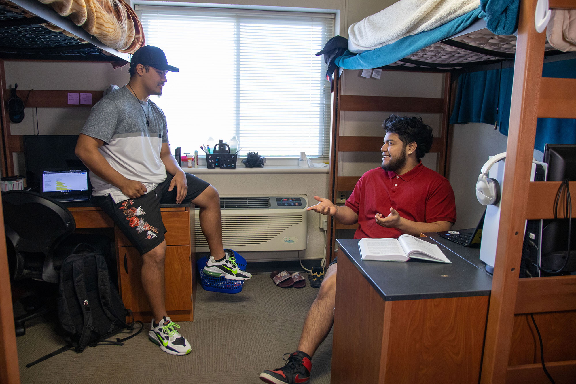 Two young men sitting on opposite sides of a dorm room under lofted beds.