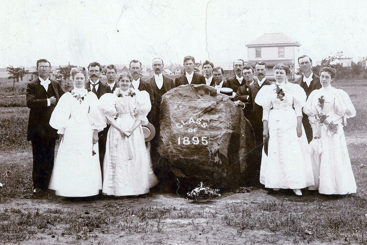 Class of 1895 included four women standing around a large rock with male students standing behind the rock. 