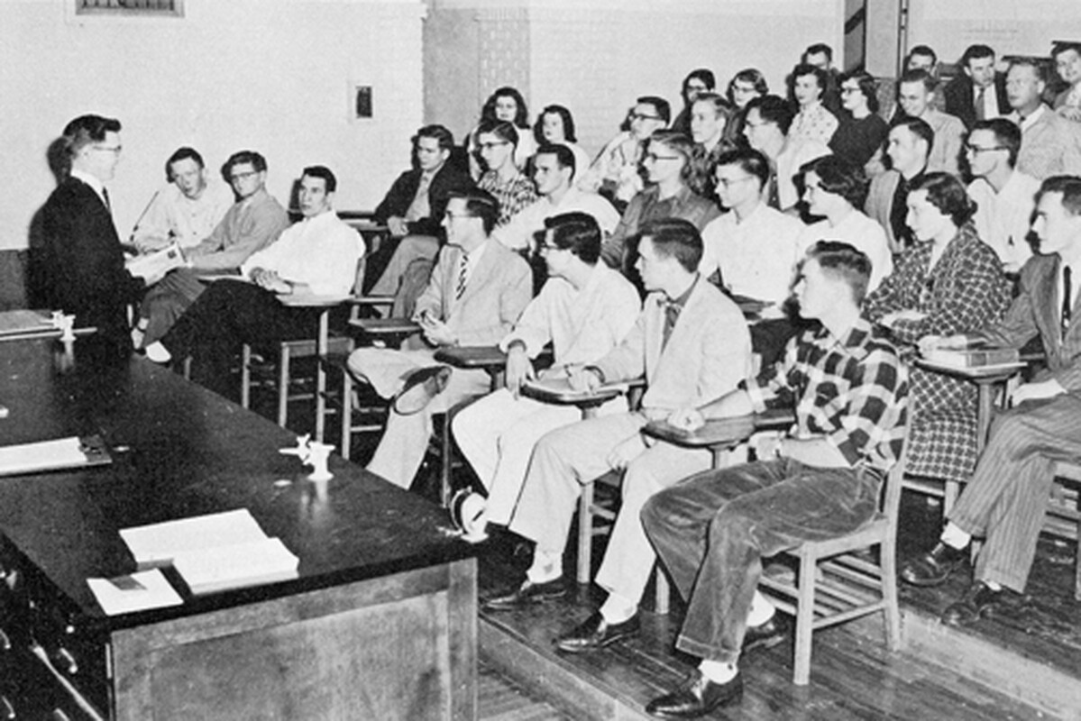 This black and white photo shows a professor on the left in front of a lecture hall with students seated at desks staggered on steps.