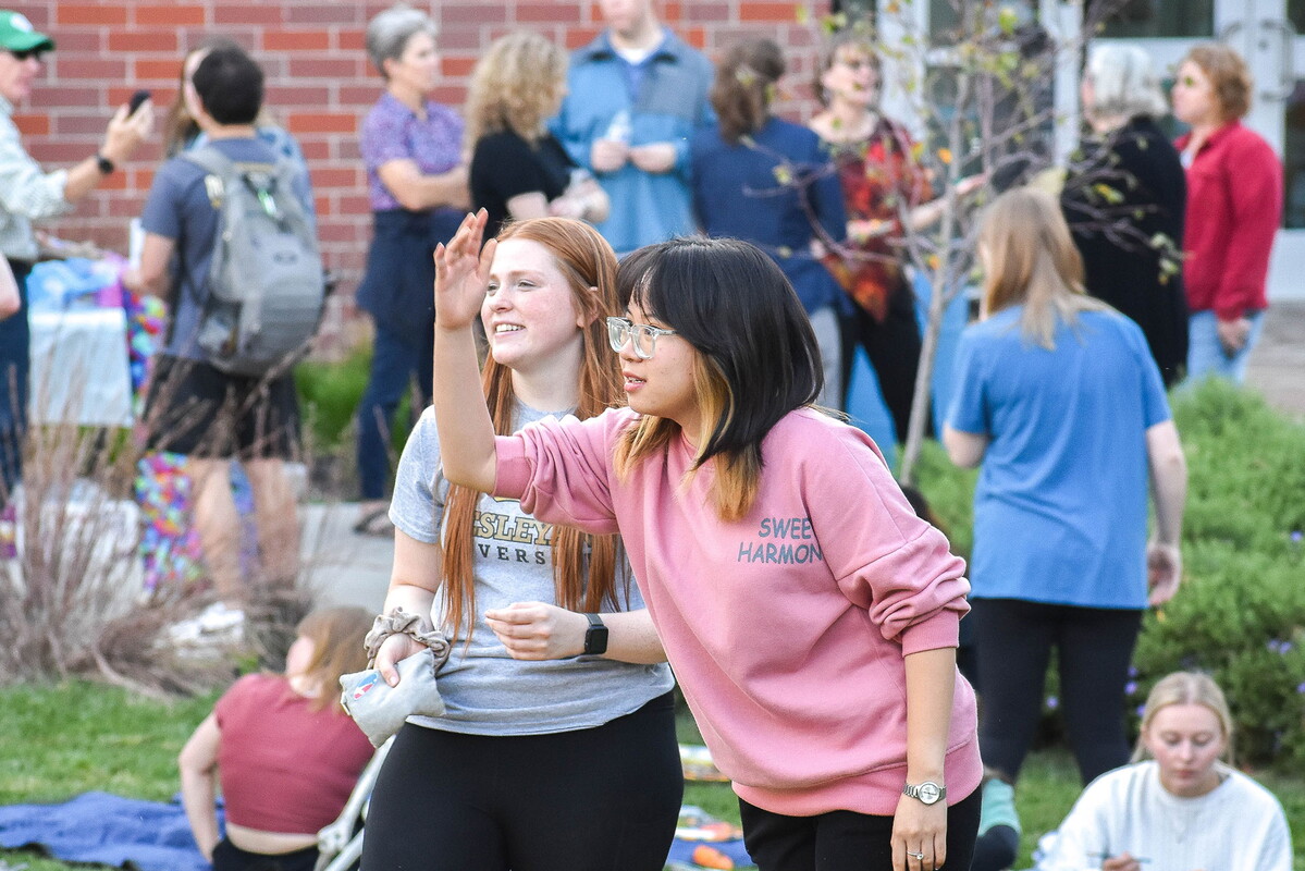 Two young women tossing a sand bag amongst many people outdoors.