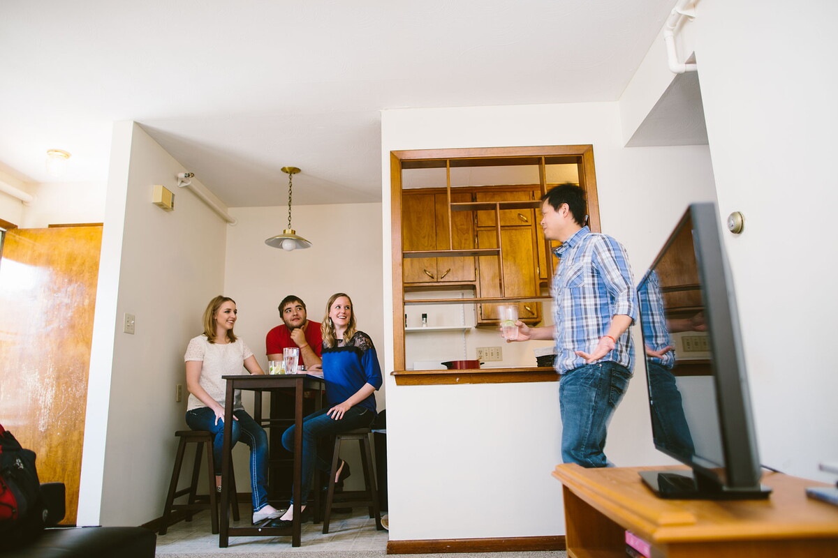 Man enter room from hallway while three people sit in the dining area of an apartment.