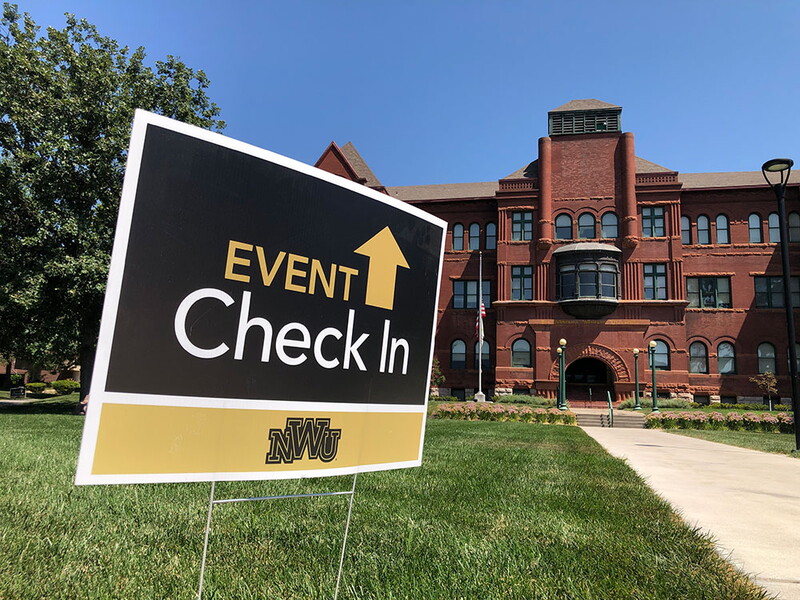 A sign staked into the ground in front of the the Old Main building reads: Event check-in with and up arrow. 
