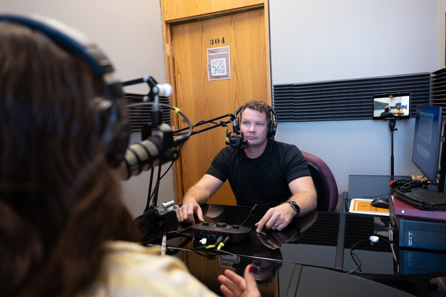 A man sits in a small room with headphones on in front of a microphone and other podcast equipment.