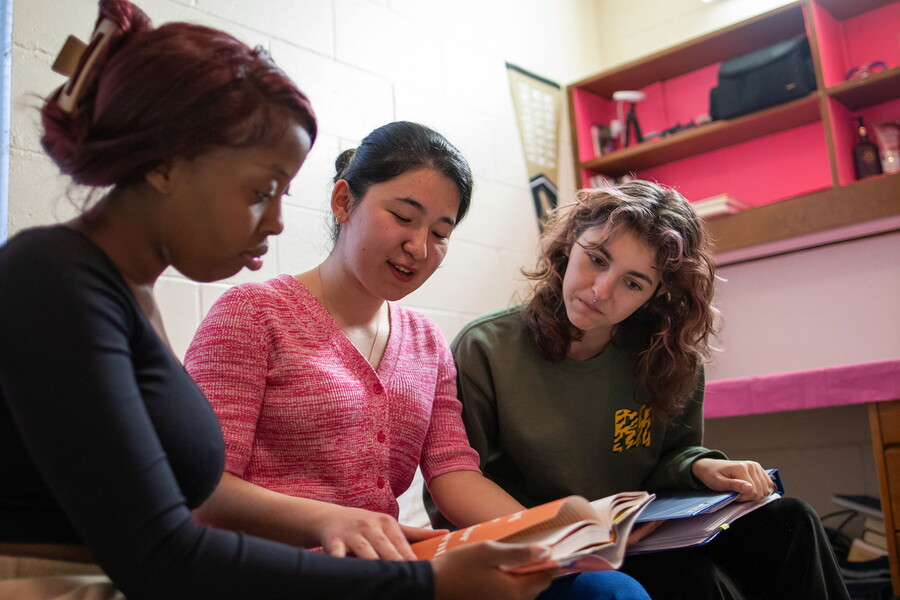 Three females looking at a book in a dorm room.