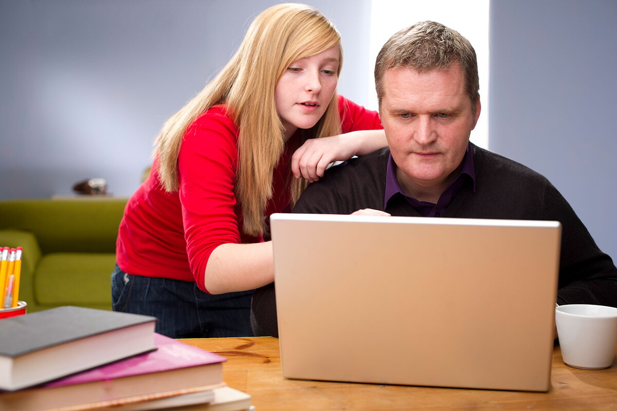 High school girl looking over her father's shoulder who is on a laptop