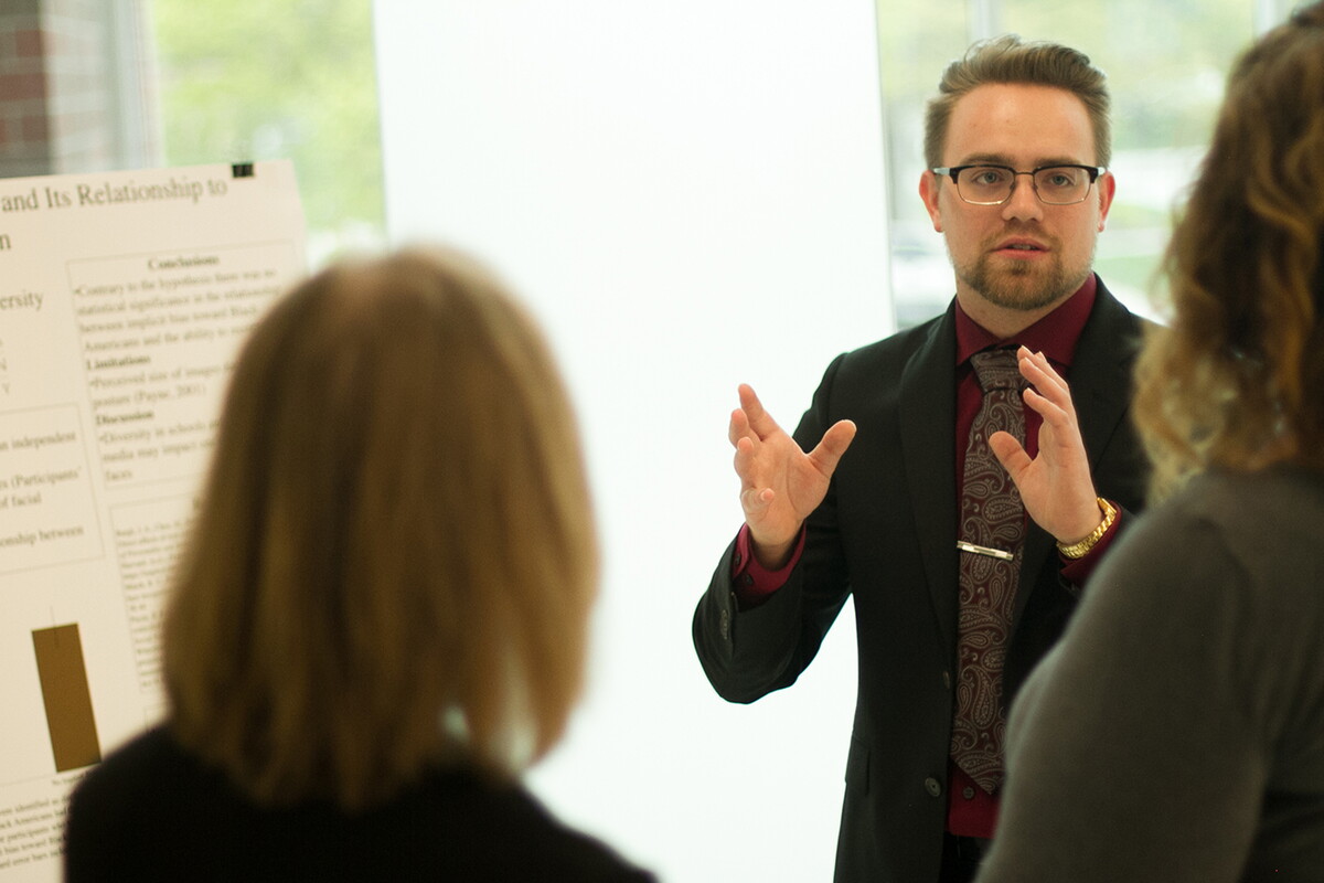 Male student in front of research poster gesturing and talking to two individuals.