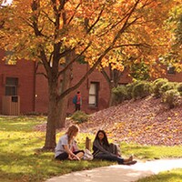 On a sunny fall day, two female students sit under a bright orange tree. 