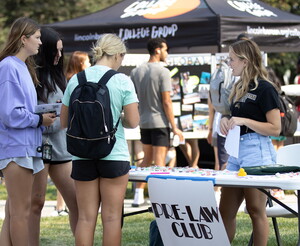 Three young women chat with young woman standing behind a table with a sign that reads Pre-law club.