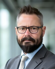 Jaye is a white man standing with his arms folded. He has short brown hair that is slightly gray along the sides and a short dark brown beard. He is wearing black framed glasses, a brown suit jacket, collared light blue dress shirt, and a light beige necktie in this formal portrait a blurred office background.