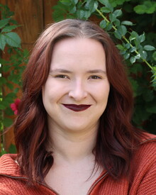 Headshot of Alex Renbarger in front of greenery covered fence. She had medium length dark reddish hair, an orange sweater, and pale skin.