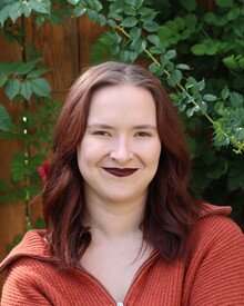 Headshot of Alex Renbarger in front of greenery covered fence. She had medium length dark reddish hair, an orange sweater, and pale skin.