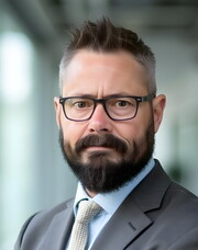Jaye is a white man standing with his arms folded. He has short brown hair that is slightly gray along the sides and a short dark brown beard. He is wearing black framed glasses, a brown suit jacket, collared light blue dress shirt, and a light beige necktie in this formal portrait a blurred office background.
