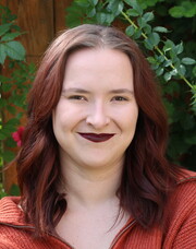 Headshot of Alex Renbarger in front of greenery covered fence. She had medium length dark reddish hair, an orange sweater, and pale skin.